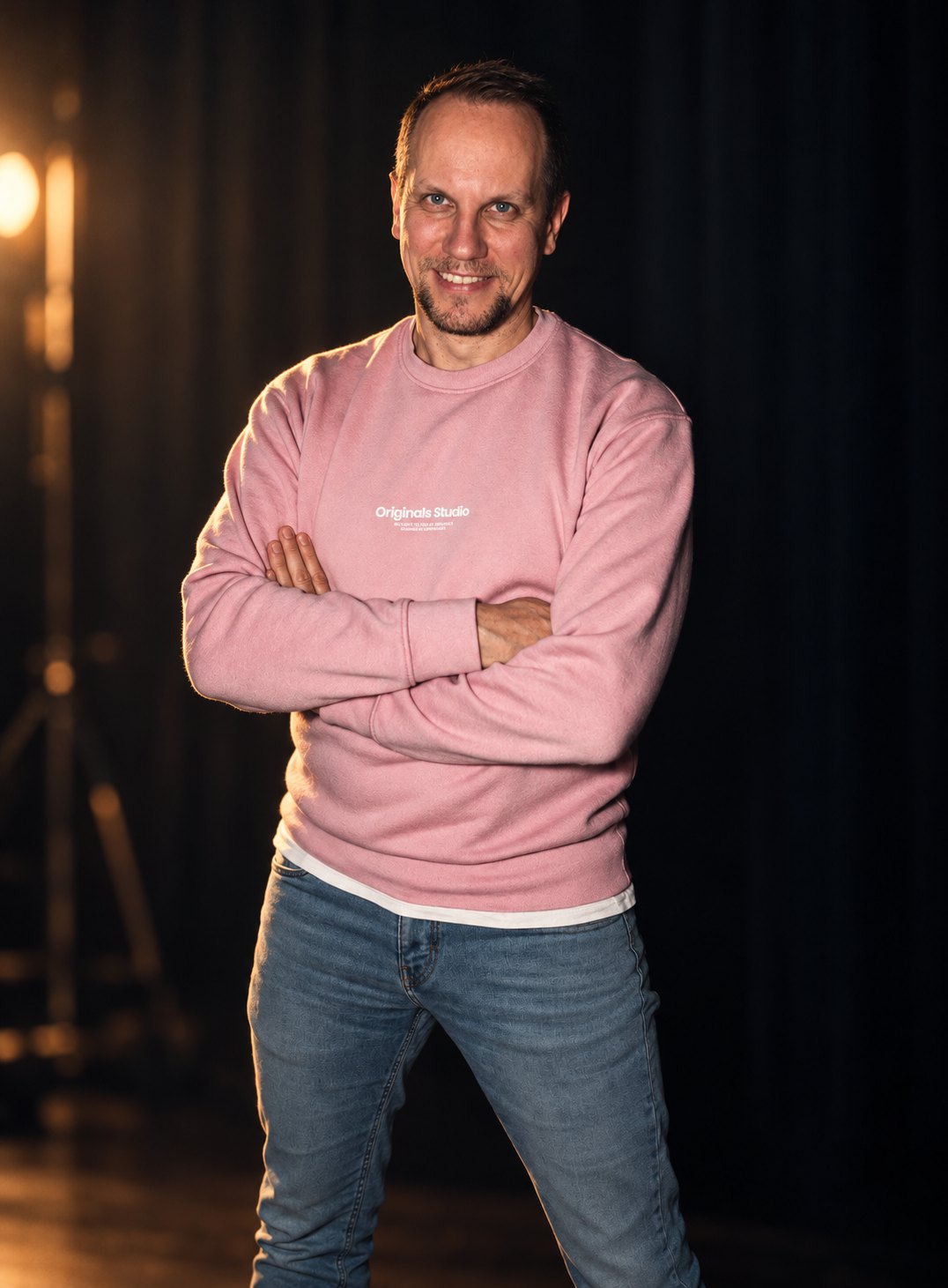 Nik Thomi mit verschränkten Armen im rosa Sweatshirt vor dunklem Bühnenvorhang